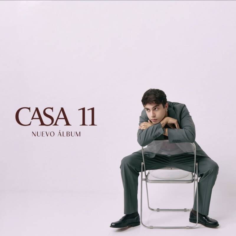 El artista Rusherking está sentado en una silla apoyando su rostro sobre el respaldo mientras viste un traje gris.